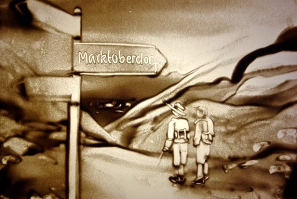 Sandbild von 2 Wanderern vor einem Wegweiser